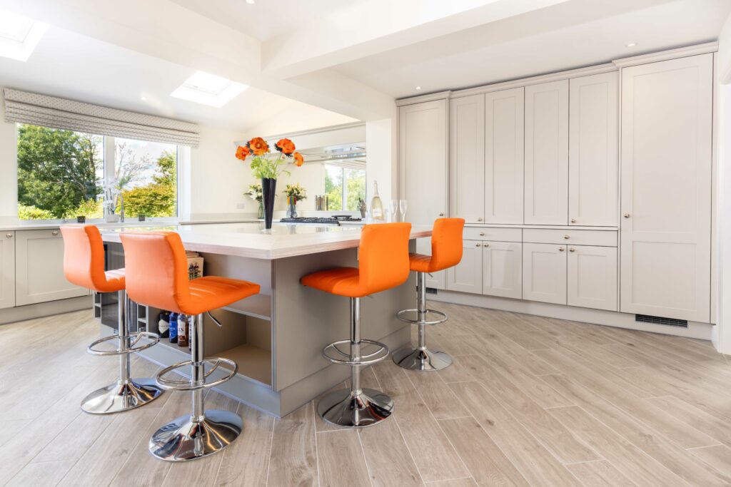 A bright, spacious kitchen with off-white cabinetry and a central island with a white countertop and orange bar stools. The floor is a pale wood, and the room is well-lit with natural light from large windows with a garden view, and recessed ceiling lights.